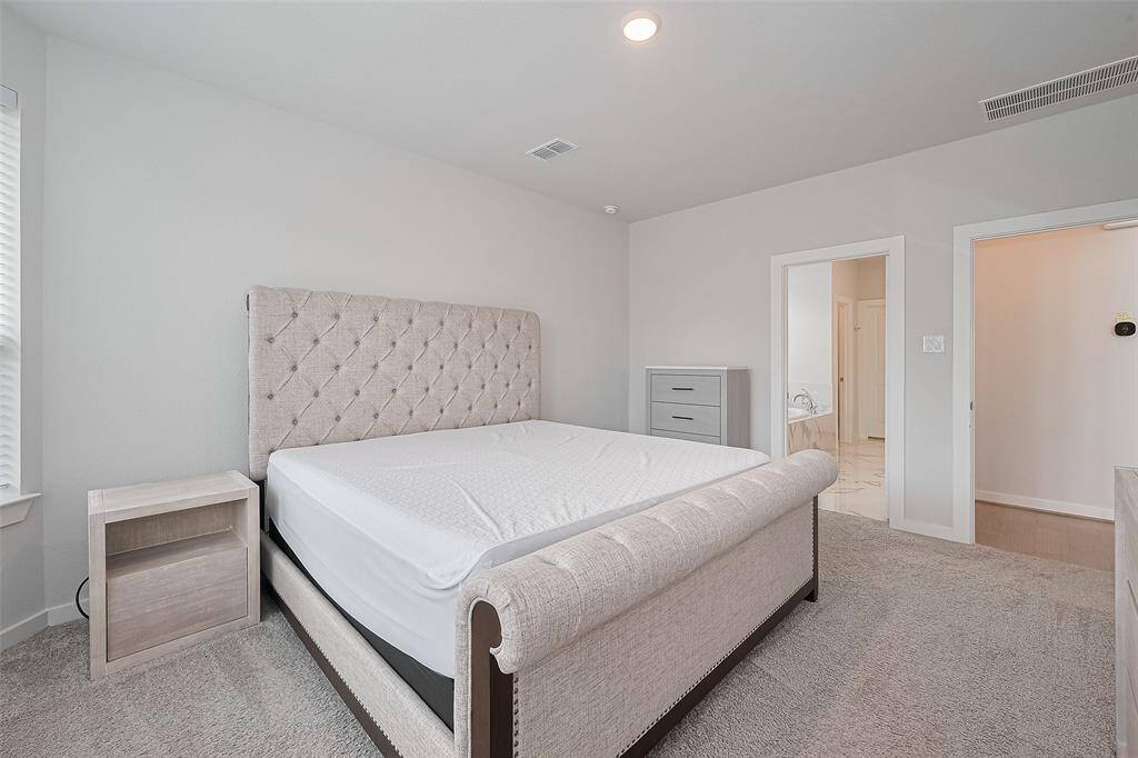 Spacious Primary bedroom with plush carpeting and neutral walls, featuring a large, upholstered bed. The room has ample natural light from a nearby window, and it includes a nightstand and dresser. There's an en-suite bathroom visible through an open door.