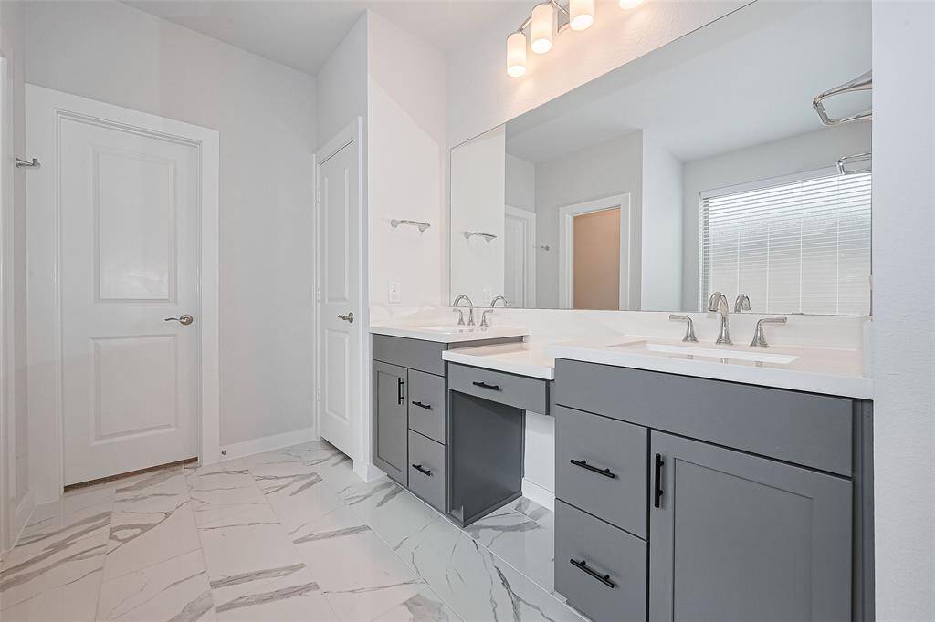 Modern bathroom features dual sinks with sleek gray cabinetry and elegant fixtures. It has a large mirror, ample lighting, and beautiful marble-like tile flooring, creating a bright and spacious feel.