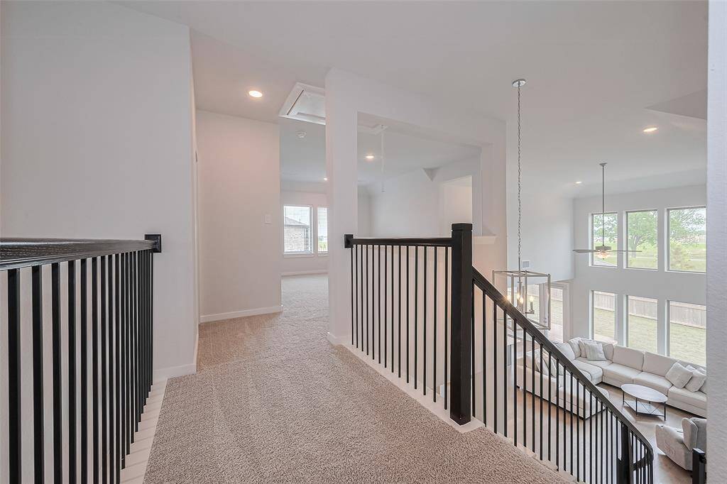 A bright, open-concept home with a modern staircase featuring sleek black railings. The upstairs landing leads to a carpeted hallway and overlooks a spacious living area with large windows, providing plenty of natural light.