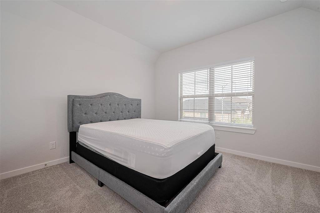 This bedroom features a plush carpet, a stylish gray upholstered bed, and a large window that allows for plenty of natural light, creating a bright and inviting space.