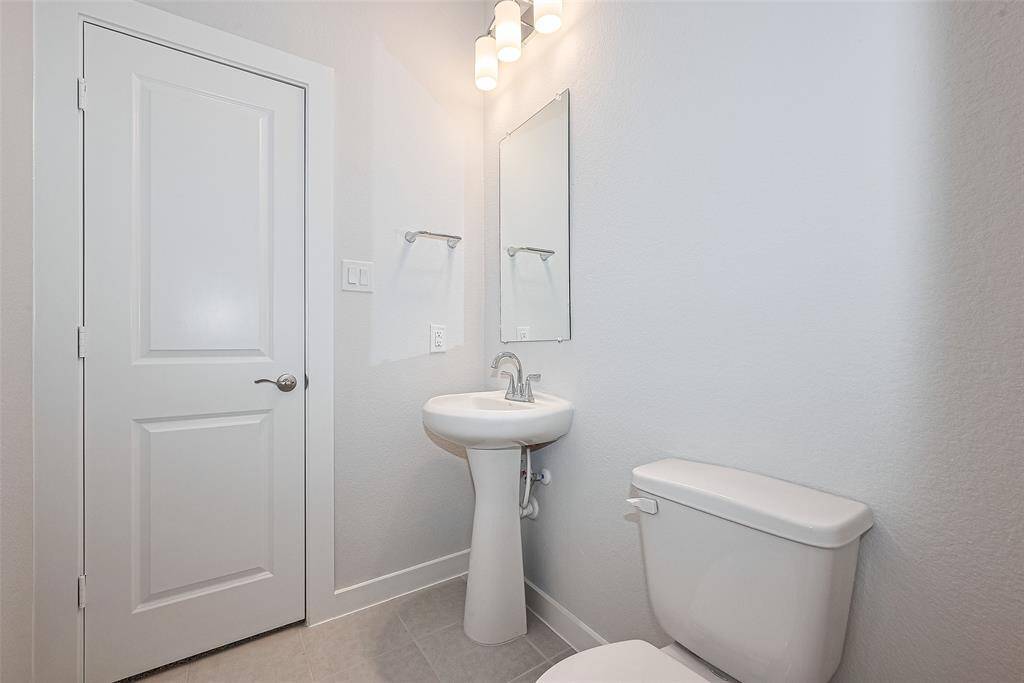 Compact, modern bathroom with a white pedestal sink, toilet, and a wall-mounted mirror. It features a three-bulb light fixture, and a door with a silver handle. The space is bright and clean, ideal for a guest or half bathroom.