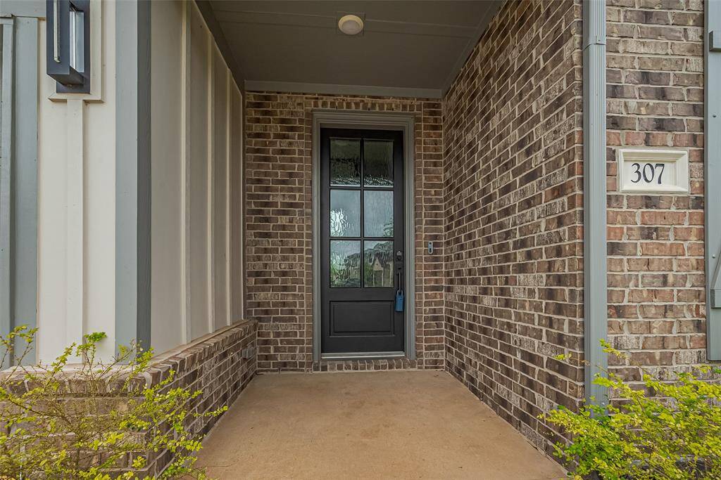 This is a welcoming front porch featuring a black door with glass panels, set against a brick exterior. The entrance is covered, providing shelter, and is complemented by modern lighting and a house number plaque.