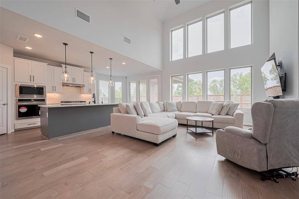 This spacious, open-concept living area features high ceilings and large windows that provide abundant natural light. The modern kitchen boasts sleek cabinetry and pendant lighting, while the cozy living space includes a comfortable sectional and a wall-mounted TV, perfect for family gatherings.