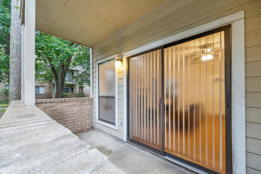 Covered patio area with a sliding glass door leading into living room. The patio is enclosed with a brick half-wall and overlooks a green, tree-filled area. Additional storage closet off of the patio.