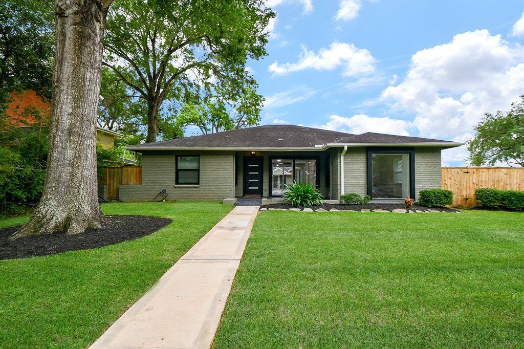 This charming single-story home features a welcoming modern facade with a dark front door, large windows, and a neatly landscaped front yard. A paved walkway leads to the entrance, and the property is surrounded by mature trees and a wooden fence, offering both curb appeal and privacy.