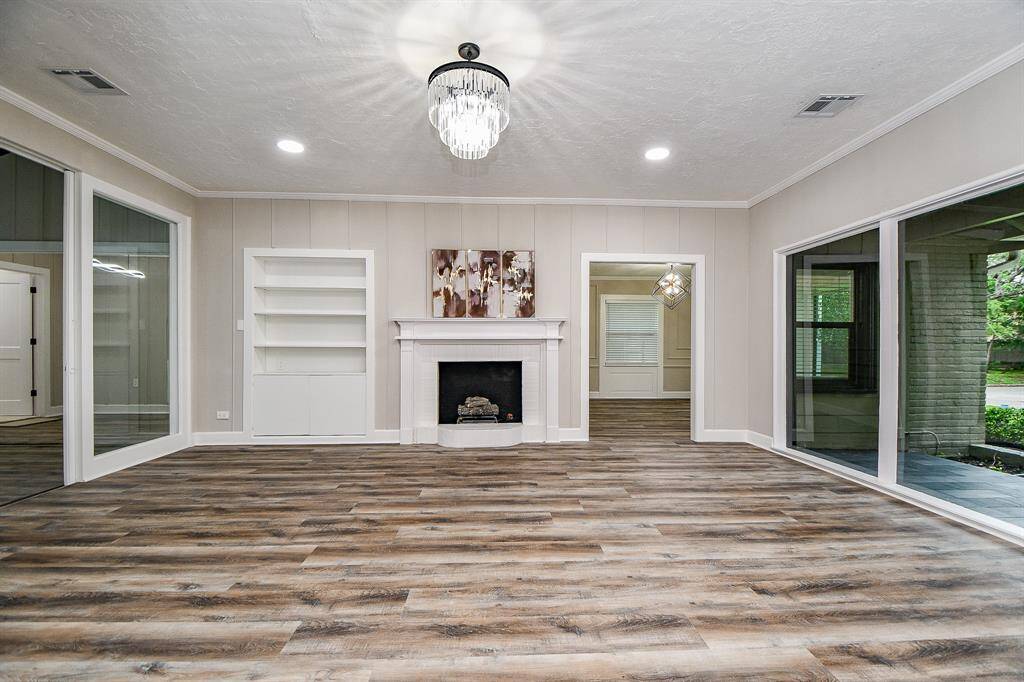 This spacious living room features elegant Laminate wood flooring, a voguish chandelier, and a cozy fireplace. Large windows provide abundant natural light, and built-in shelving offers convenient storage. The neutral color palette enhances the room's inviting atmosphere.