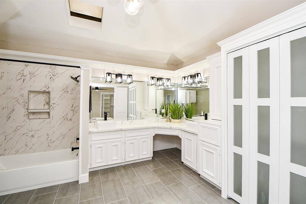 Primary modern bathroom ensuite features elegant marble-tiled walls, a spacious Quartz vanity with dual sinks, and contemporary fixtures. The large mirrors and stylish lighting enhance the room's brightness, with ample cabinet space and view of closet #3. Also a sleek new bathtub, fixtures and gray floor tiles complete the sophisticated look.