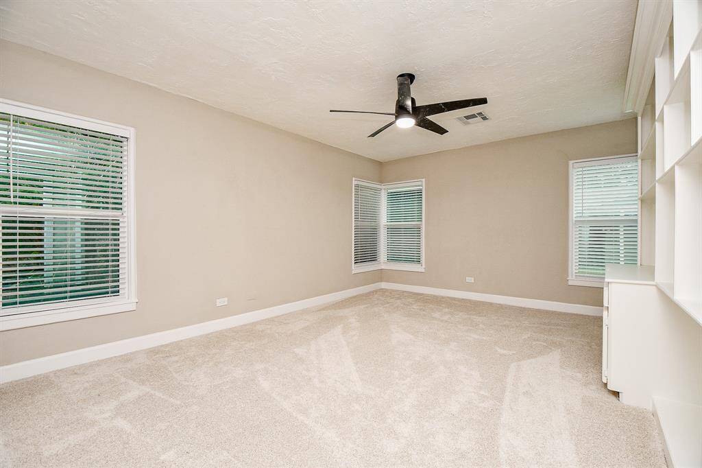 This spacious Secondary bedroom -2 features neutral walls, plush carpeting, and ample natural light from multiple windows. It includes a modern ceiling fan and built-in shelving, offering a cozy and functional space.