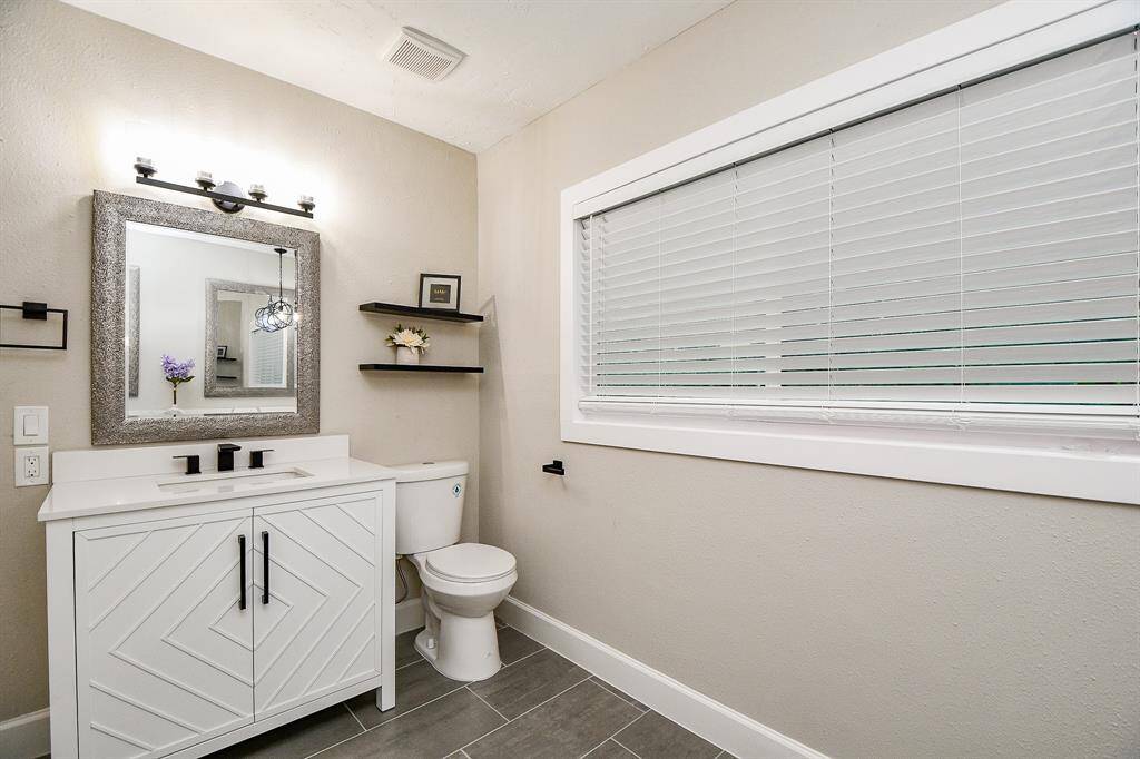 This modern bathroom with neutral tones, featuring a sleek vanity with a geometric design, a large framed mirror, and black fixtures. It has open shelving for decor, and light gray tile flooring.