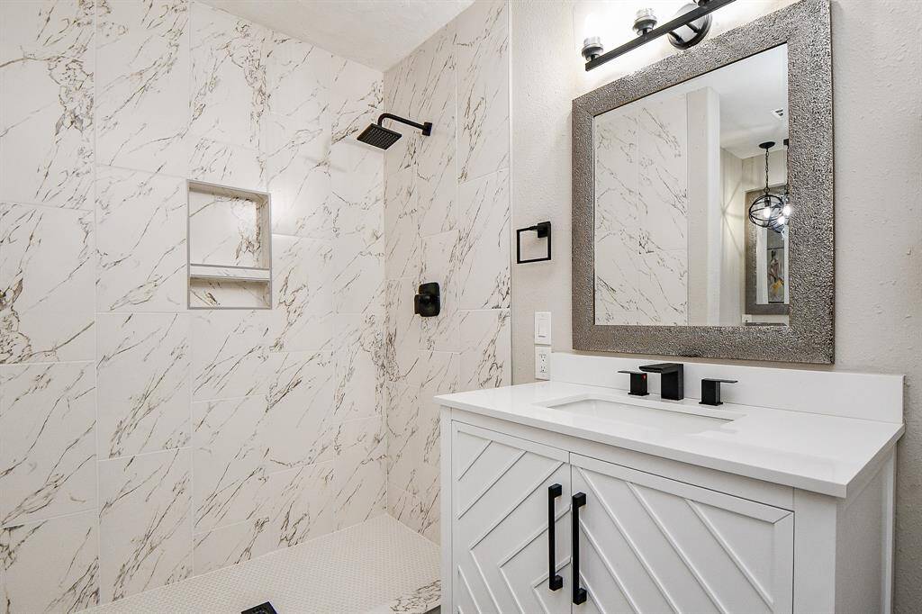 Modern hall bathroom Pic#2, features elegant marble-like tile walls with a spacious walk-in shower. The vanity has a sleek white countertop, black fixtures, and a large decorative mirror, creating a stylish and contemporary look.