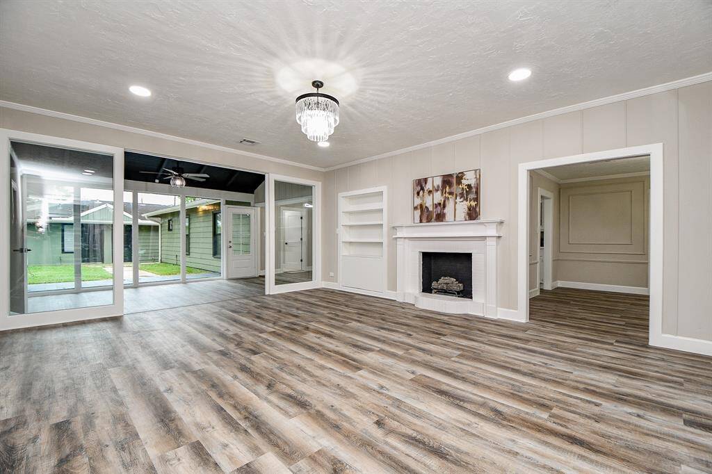 This spacious Family Room features elegant laminate wood flooring, a cozy fireplace with built-in shelving, and abundant natural light from large glass doors leading to a sunroom and outdoor space. The neutral color palette and modern chandelier enhance the room's inviting atmosphere.