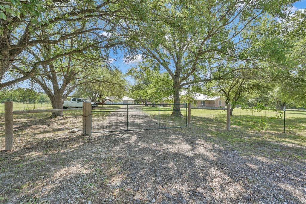 A spacious property with a gated entrance and gravel driveway. The area is shaded by large trees, providing a serene, rural atmosphere.