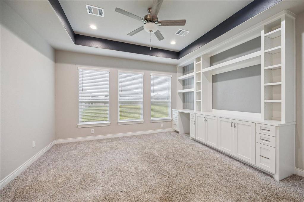 This room features a tray ceiling with a ceiling fan, abundant natural light from three large windows, built-in white cabinetry and shelving, ideal for storage or a home office setup.