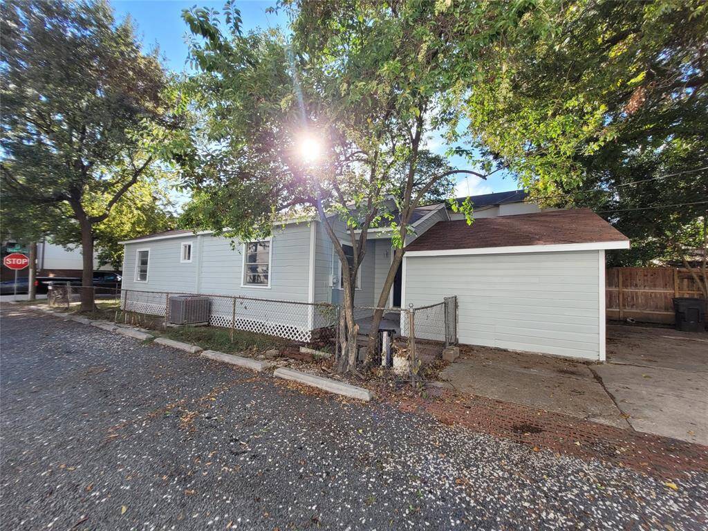 Quaint single-story home with light siding, nestled among mature trees. Features a small fenced area and adjacent parking space. Located on a quiet street corner with a stop sign visible.