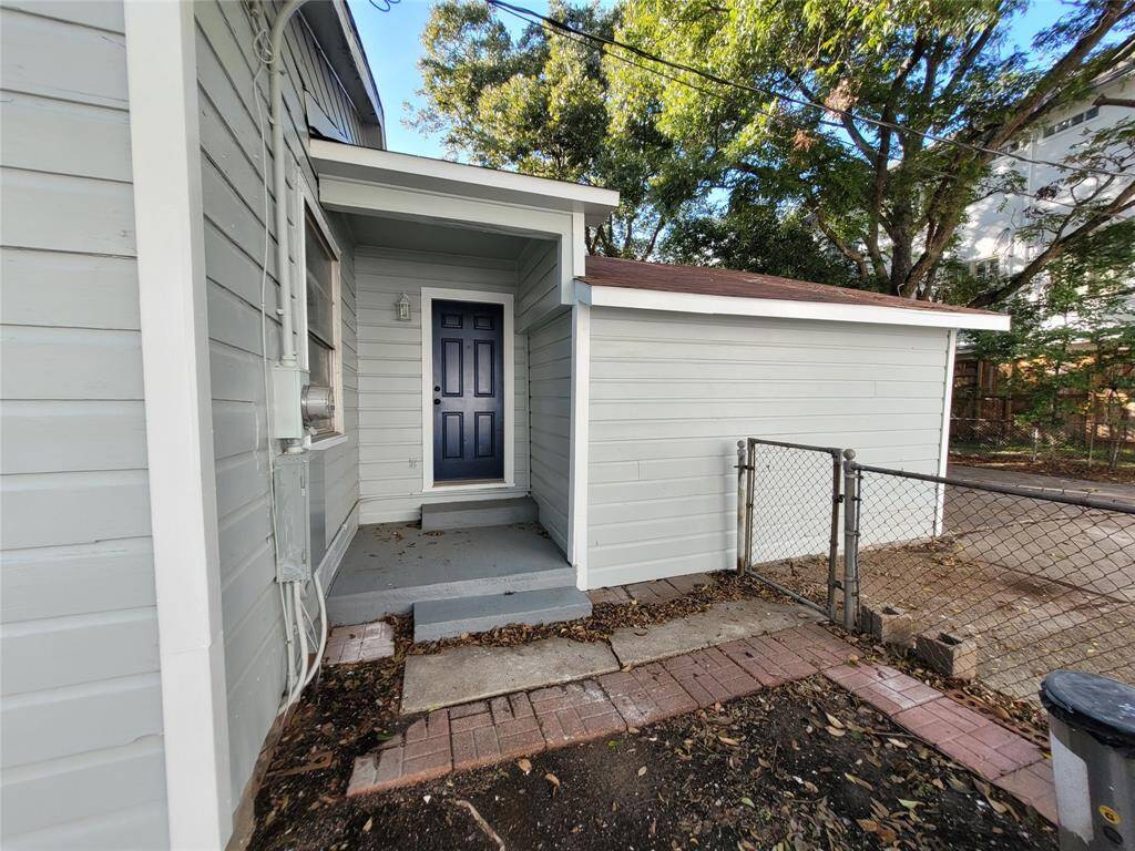 This photo shows a side entrance to a house with light gray siding and a dark blue door. It features a small concrete step and a brick path, with a fenced area and trees in the background, offering privacy and a cozy feel.