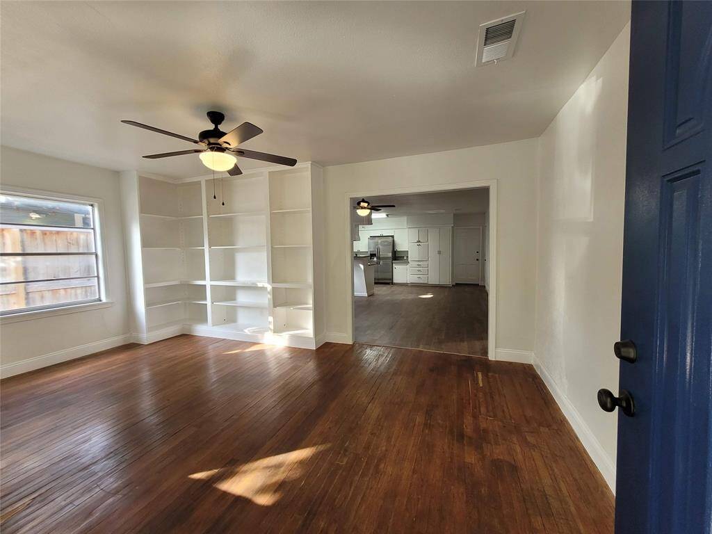 Bright, open living space with hardwood floors, built-in shelves, and a ceiling fan. The room flows into a modern open concept kitchen with stainless steel appliances, creating a welcoming and functional home environment.