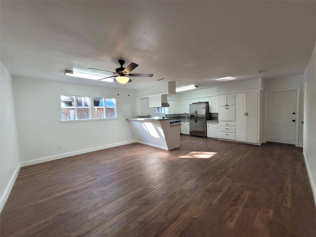 This open-concept living space features hardwood floors, a modern kitchen with stainless steel appliances, white cabinetry, and a ceiling fan. Large windows provide ample natural light.