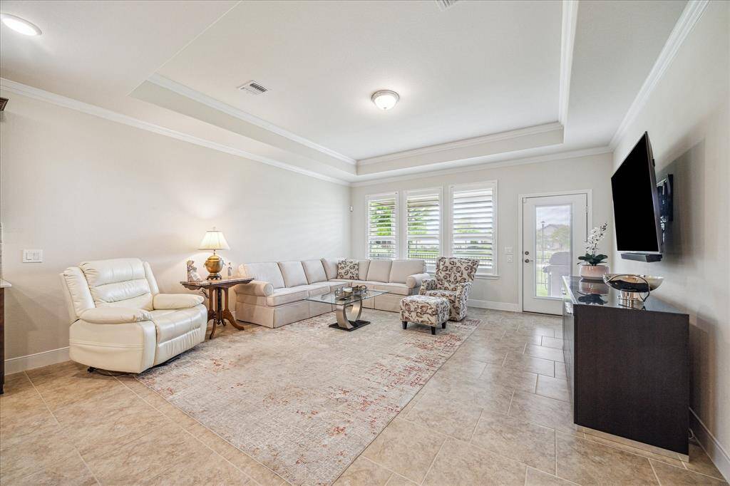Bright and spacious living room with a cozy sectional sofa and armchair, large windows with shutters, tiled flooring, and a wall-mounted TV. The room features neutral tones and opens to an outdoor area through a glass door.