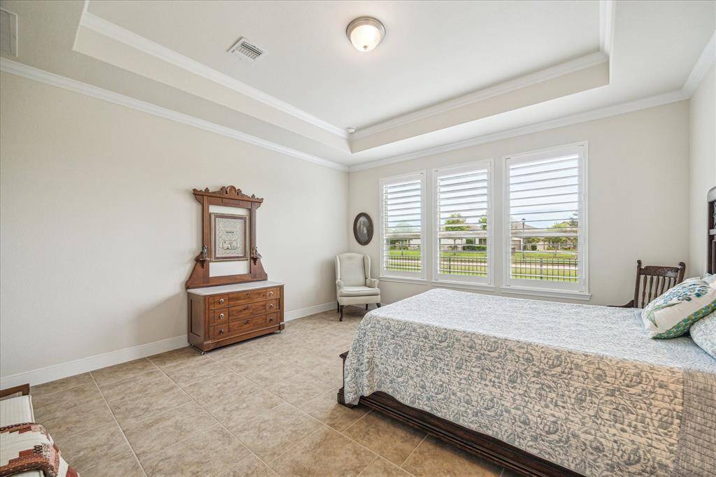 Bright and spacious bedroom with large windows, neutral walls, and tiled flooring. Features a decorative ceiling, classic wooden furniture, and a comfortable armchair, offering a cozy and elegant atmosphere.