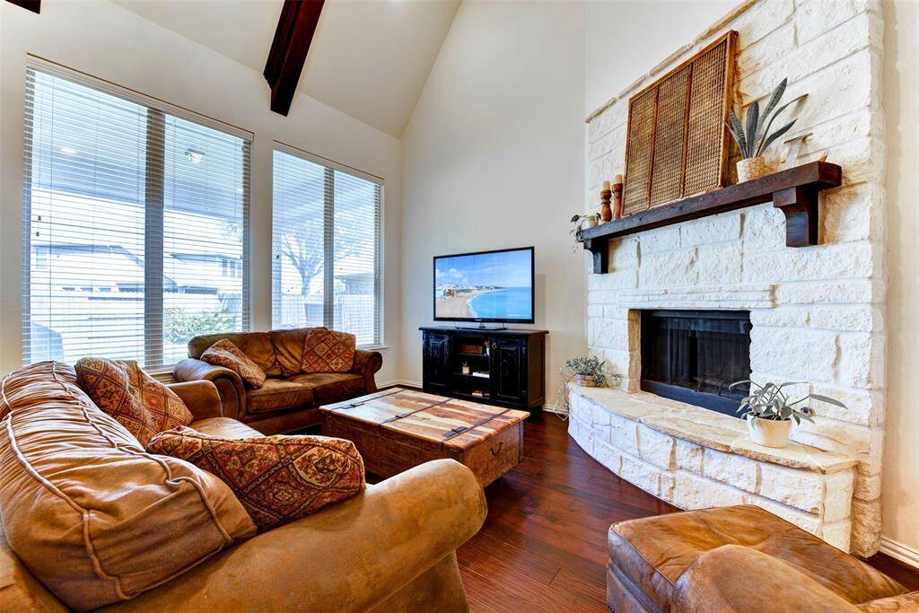 This cozy living room features high vaulted ceilings with wooden beams, a stone fireplace, and large windows letting in ample natural light. The hardwood floors and comfortably spaced sitting area create an inviting space perfect for relaxation