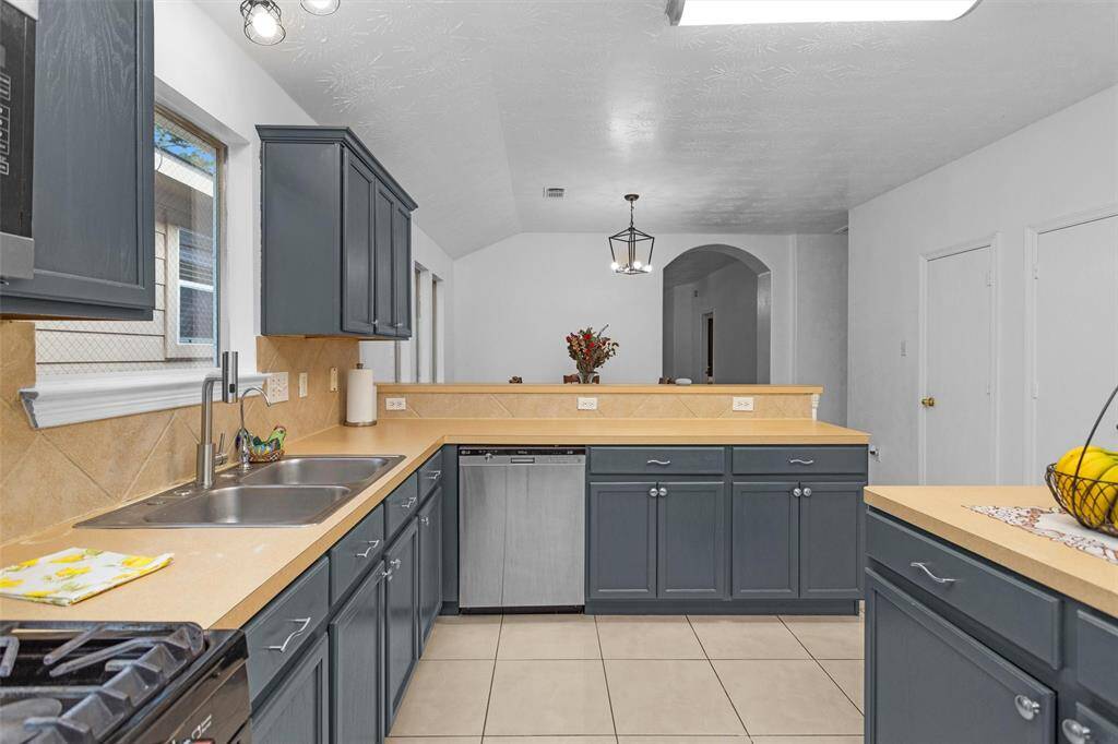Modern kitchen with teal cabinets, stainless steel appliances, and a double sink. The space features a beige countertop and tile backsplash, with a view into the dining area through an open archway. Bright and inviting with overhead lighting and natural light from a window.