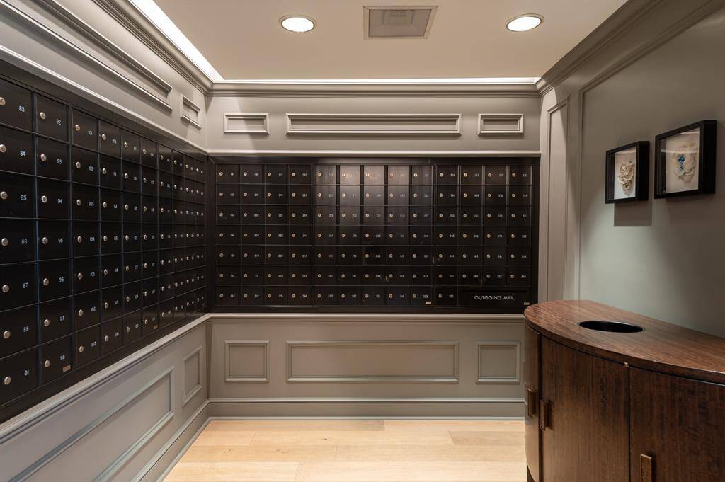 This spacious living room, carpeted living area with neutral walls, featuring a large mirrors on one side. It opens into a kitchen with white cabinetry and a visible entrance to another room. The space is well-lit with ceiling fixtures.