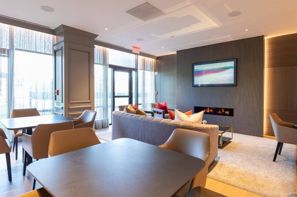 This elegant public sitting room features a large window with sheer curtains, allowing natural light to illuminate the space. It includes a modern gray sofa with vibrant red and blue cushions, a stylish glass coffee table, and contemporary art on the wall. The room is accented with a sleek gold lamp and plush gray armchairs, creating a sophisticated and inviting atmosphere.