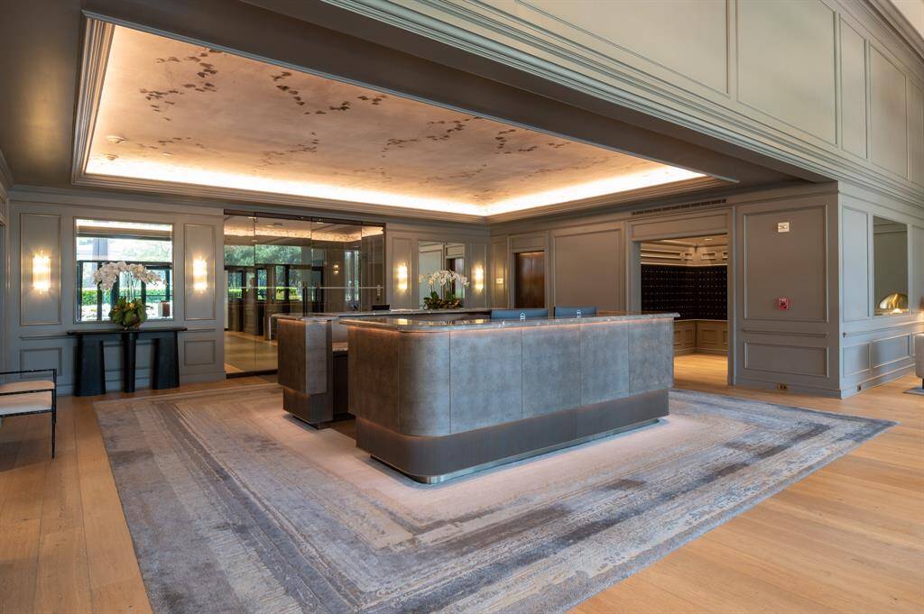 This elegant sharing living room features high ceilings, a large chandelier, and stylish wall paneling. It includes a comfortable seating area with a plush sofa, armchair, and a glass coffee table on a soft rug. Large windows bring in plenty of natural light, enhancing the sophisticated decor.