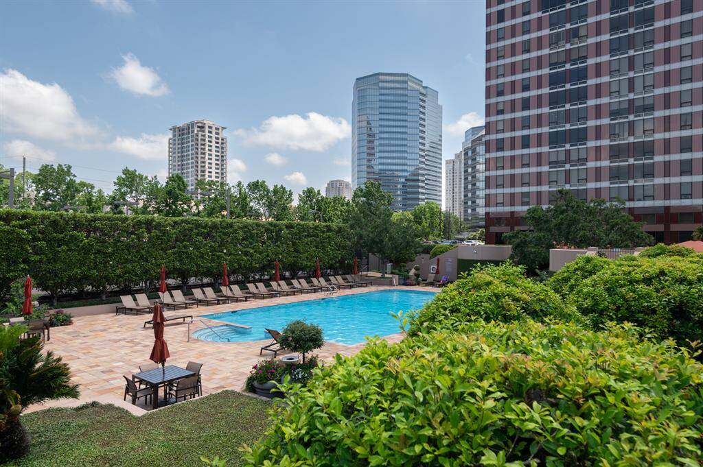 This photo showcases a modern apartment complex with a spacious outdoor pool area, surrounded by lush greenery and ample lounge seating. The setting offers a relaxing oasis amidst a cityscape of tall buildings, perfect for enjoying sunny days.