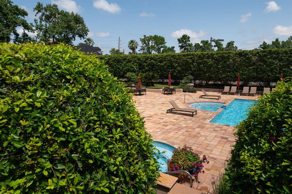 This photo showcases a backyard with a spacious patio area featuring a swimming pool and hot tub. The area is surrounded by lush greenery and includes lounge chairs and patio tables with red umbrellas, creating a private and inviting outdoor space.