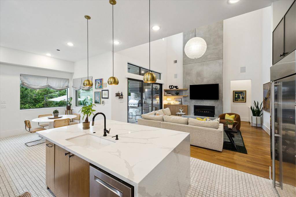 This stunning open-concept kitchen and family room showcases seamless modern design, highlighted by a dramatic waterfall-edge island, brass pendant lighting, and soaring ceilings that enhance the sense of space and light. The sleek kitchen transitions effortlessly into the inviting living area, where a contemporary fireplace and statement feature wall create a warm, stylish focal point for relaxed