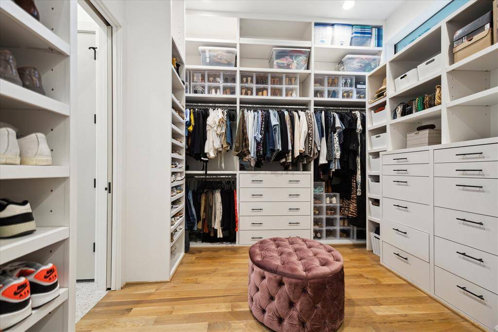 This expansive primary closet is a luxurious blend of style and organization, featuring custom built-ins, dedicated shoe storage, and abundant drawer space, all set against the warmth of wide-plank hardwood floors. A tufted velvet ottoman anchors the room, offering both elegance and functionality in this beautifully curated dressing retreat.