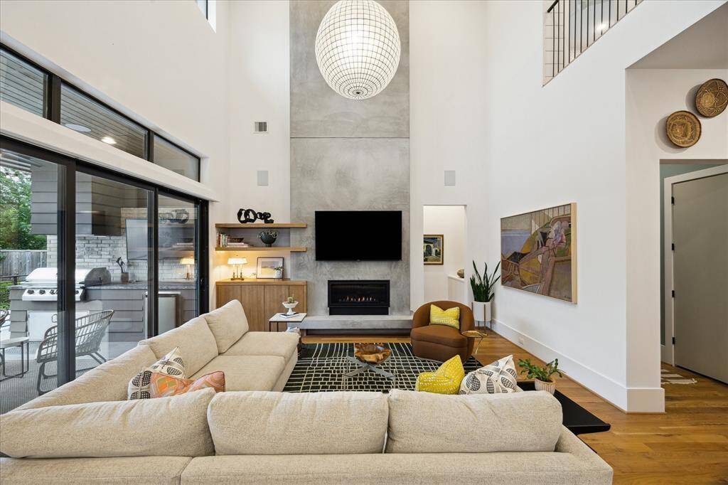 Soaring ceilings and expansive glass sliders flood this striking living room with natural light, highlighting the sleek concrete fireplace as a bold focal point. Warm wood floors and curated design accents create an inviting balance of modern sophistication and comfortable elegance.