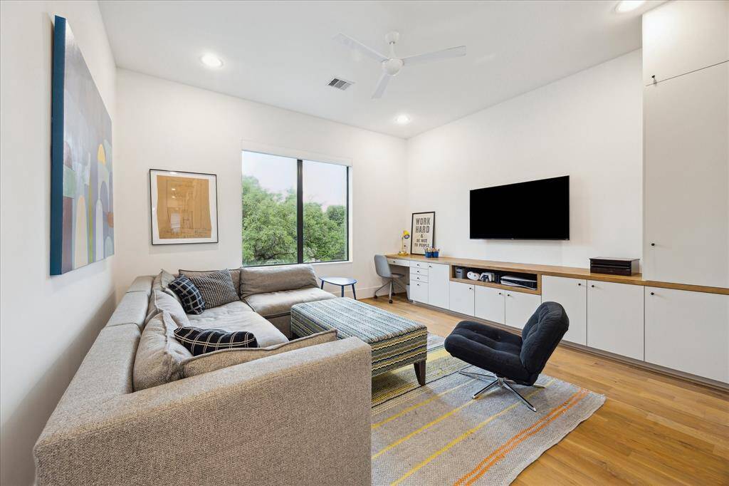 This expansive second-floor game room offers a stylish and versatile retreat, featuring sleek built-in cabinetry, a dedicated workspace, and ample room for lounging or entertaining. Bathed in natural light from a large picture window and finished with warm hardwood flooring and contemporary furnishings, the space balances comfort and function with modern flair.