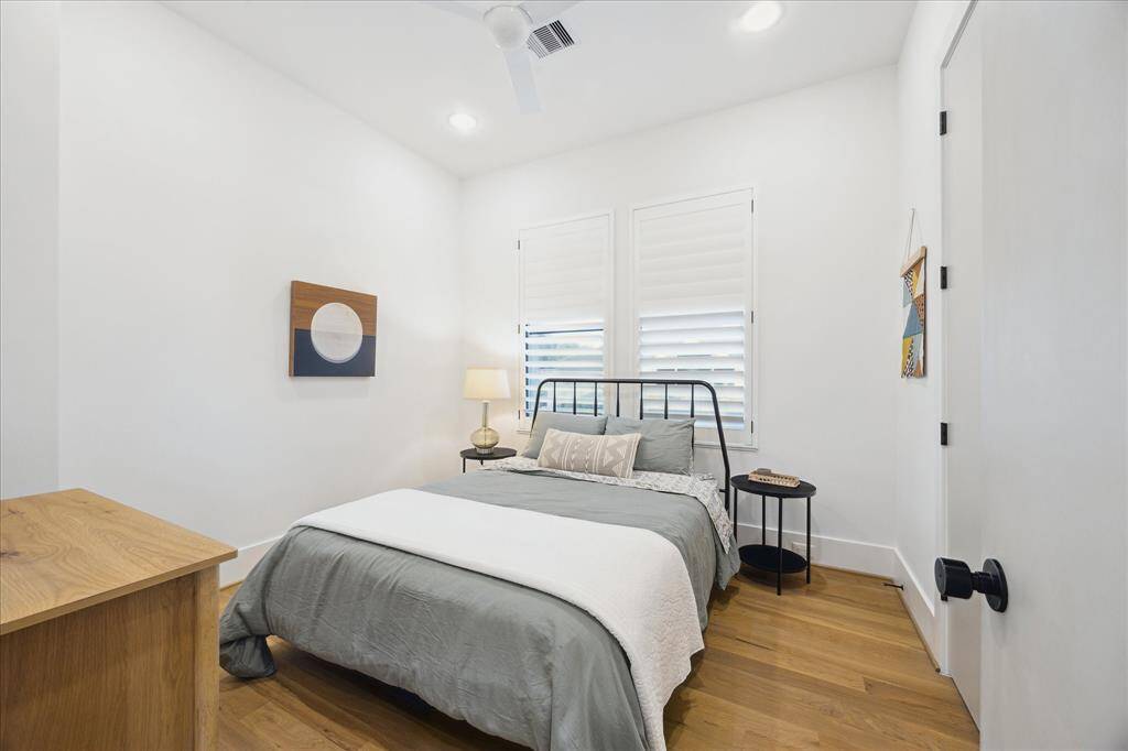 This serene secondary bedroom is grounded by warm hardwood floors and bathed in soft natural light, with crisp white walls, modern window treatments, and minimalist furnishings that evoke a sense of calm and simplicity.