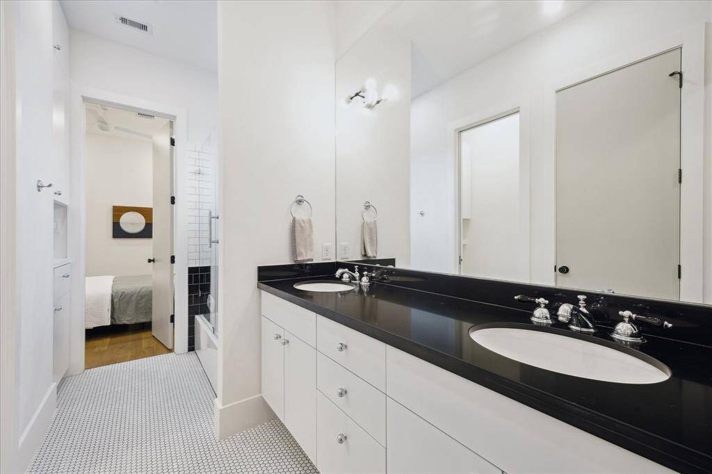 This sleek secondary bathroom pairs classic black-and-white finishes with modern functionality, featuring dual sinks with polished black countertops, crisp white cabinetry, and chic penny tile flooring. A glass-enclosed tub and shower combo with stylish subway tile offers a timeless and practical design for shared use.