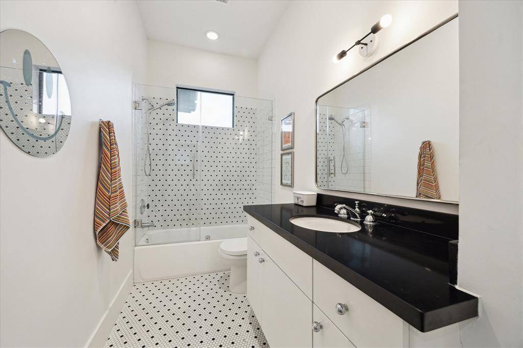 This secondary bathroom combines classic charm with modern touches, featuring bold black countertops, crisp white cabinetry, and playful penny tile flooring. The tub and shower are framed by a glass enclosure and accented with timeless mosaic tilework, while high ceilings and a transom window invite in natural light for a fresh, airy feel.