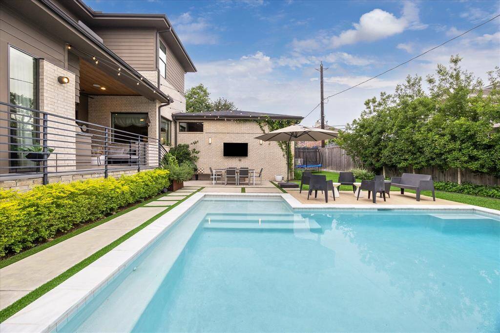 This thoughtfully designed backyard showcases a sparkling pool surrounded by lush landscaping, a stylish sundeck with modern lounge seating, and a covered outdoor dining area anchored by a wall-mounted TV—creating a resort-style atmosphere perfect for relaxation and entertaining.