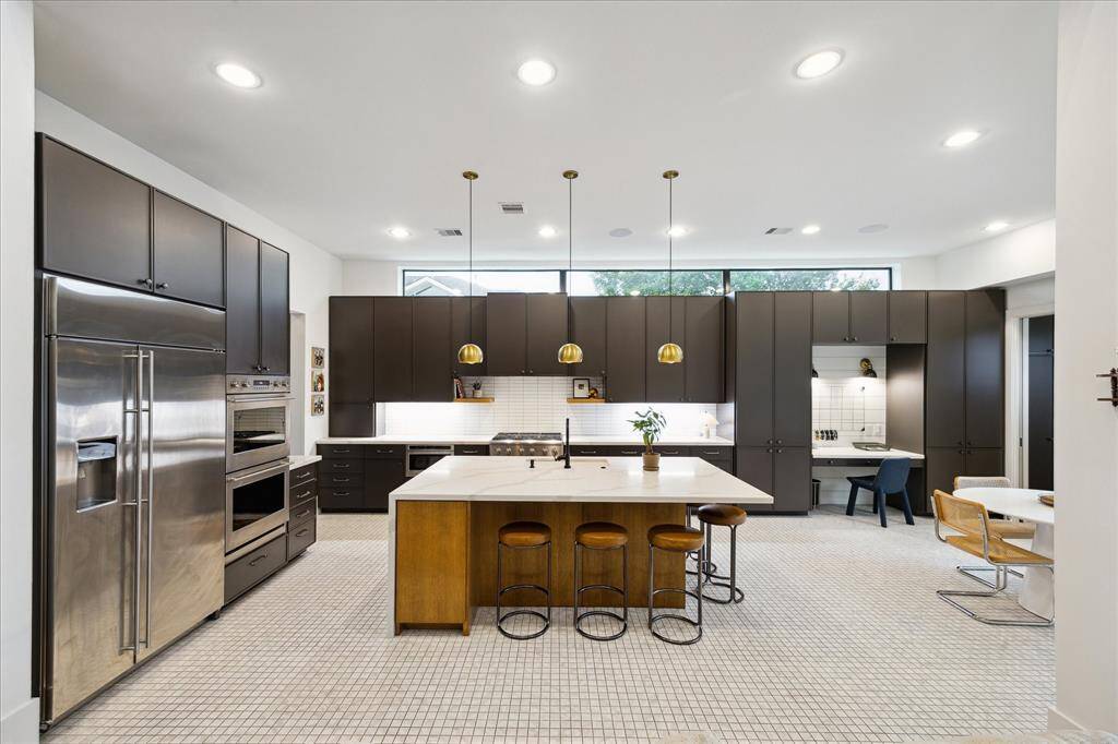 This designer kitchen exudes modern elegance with sleek matte cabinetry, striking brass pendant lighting, and a waterfall-edge island that anchors the space with warmth and functionality. Expansive upper transom windows invite natural light, while the integrated workspace and premium appliances cater to both everyday living and elevated entertaining.
