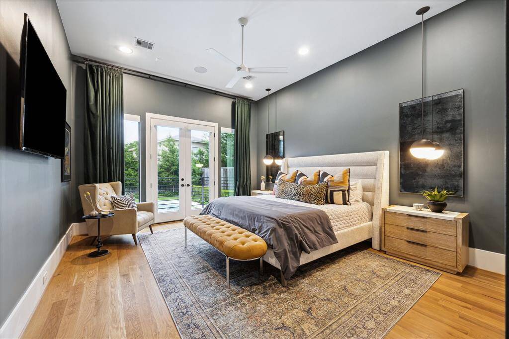 The primary suite is a refined sanctuary, artfully designed with rich hardwood floors, dramatic accent walls, and soaring ceilings that amplify its serene ambiance. Elegant pendant lighting, luxe textiles, and an upholstered bed frame create a polished yet inviting retreat, while French doors open to the outdoor oasis, seamlessly blending indoor comfort with tranquil views.