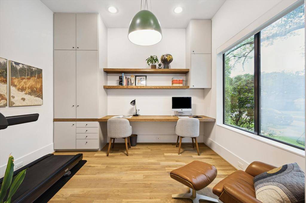 Bathed in natural light from a picture window, this thoughtfully designed home office features custom built-ins, floating shelves, and a generous dual workstation for effortless productivity. Warm wood floors and modern finishes create a serene, stylish environment ideal for focused work or creative inspiration.