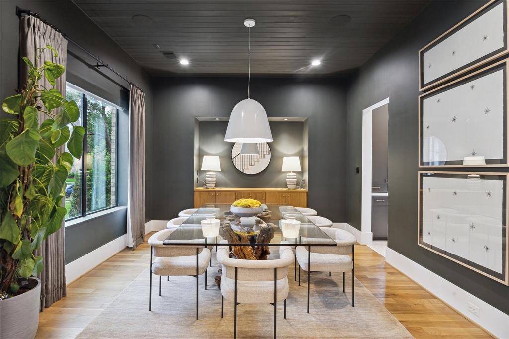 Sophisticated and moody, this formal dining room stuns with dramatic dark walls, a sleek glass-top table anchored by a sculptural wood base, and layered lighting that creates an inviting ambiance. The built-in sideboard and curated artwork elevate the space, blending modern design with effortless elegance.