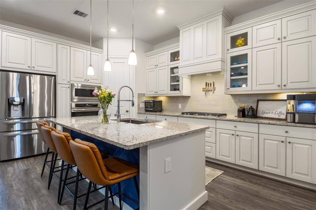 This jaw dropping modern kitchen features beautiful white cabinetry, walk-in pantry, a central island with custom blue paneling underneath and granite countertops. Pendant lighting and ample storage add to its functionality, complemented by a stylish backsplash and hardwood flooring.