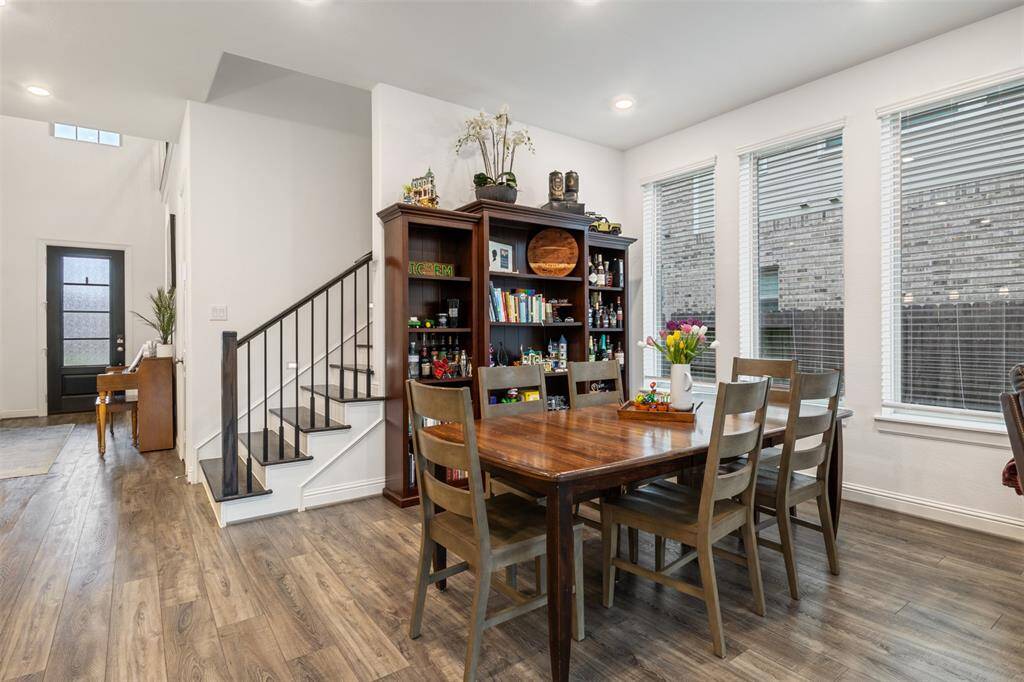 The inviting dining area in the heart of the home features natural light from large windows, and elegant vinyl flooring. The adjacent staircase leads to an open entryway with a modern door, creating a warm and functional living space.