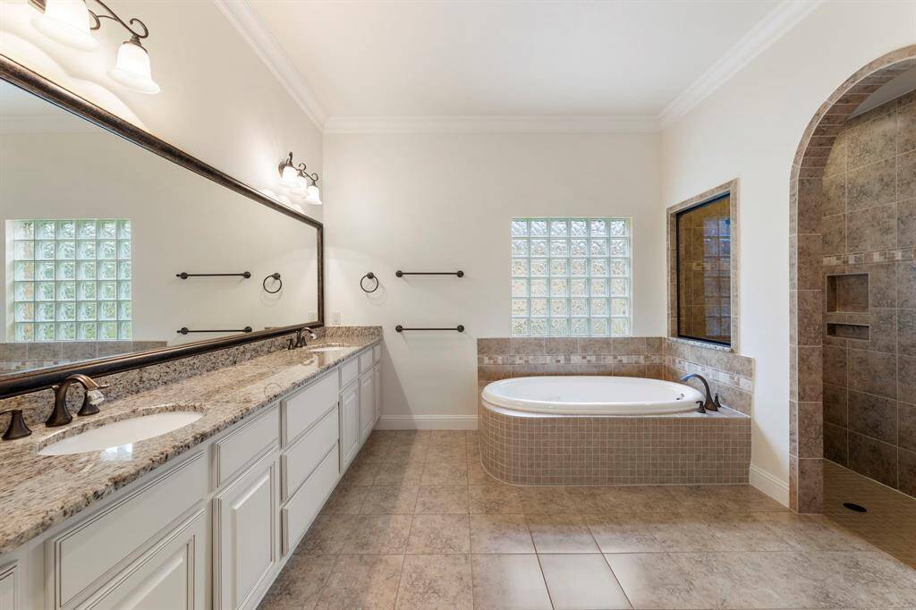 The luxurious primary bath is a true retreat, complete with an oversized dual-sink vanity, deep soaking tub framed by glass block windows, and a walk-in shower with elegant tilework and built-in shelving.