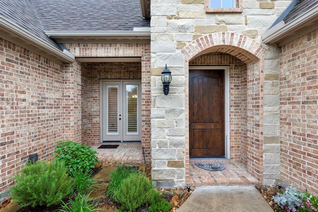 The brick-lined entry features a rich wood front door and double patio doors, framed by lush landscaping and natural stone accents for a warm, welcoming first impression.