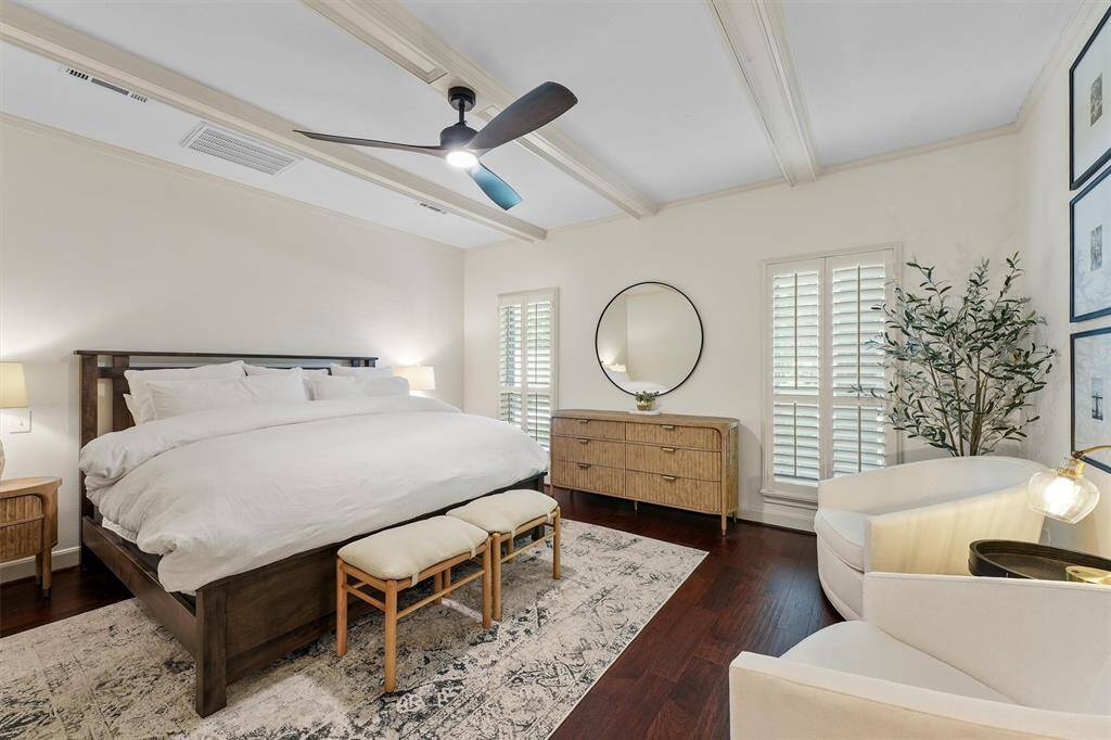 First floor primary bedroom is inviting and elegant, featuring dark wood flooring, and a stylish ceiling fan. Room for a cozy seating area, dresser, and ample natural light through shuttered windows complete the serene space.