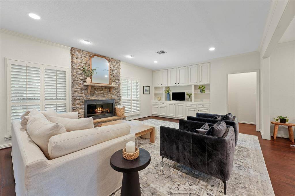This cozy living room features a stone fireplace, built-in cabinetry, and a mix of plush seating. It has a bright, airy feel with white walls, large windows with shutters, and recessed lighting. Ideal for relaxing and entertaining.