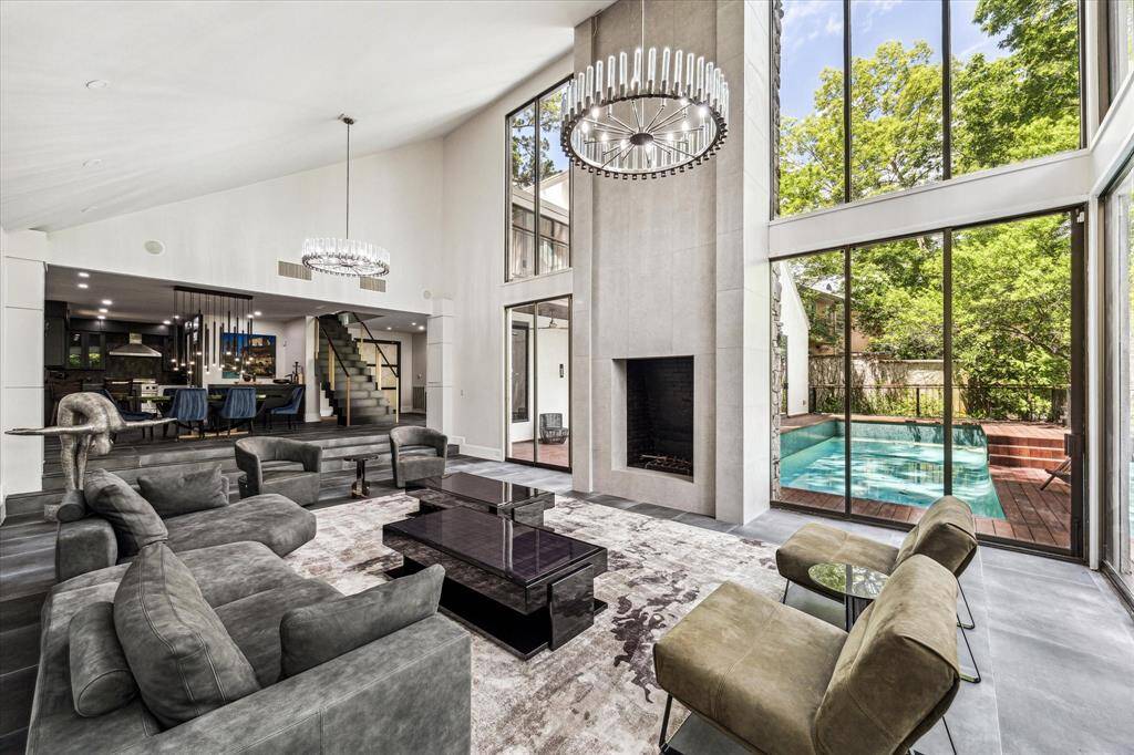 luxurious open concept living room boasting soaring ceilings, a dazzling chandelier, wide windows that flood the space with natural light, and a magnificent stone fireplace as its focal point. The grandeur of the high ceilings amplifies the sense of space and airiness, while the chandelier adds a touch of sophistication and warmth to the room.