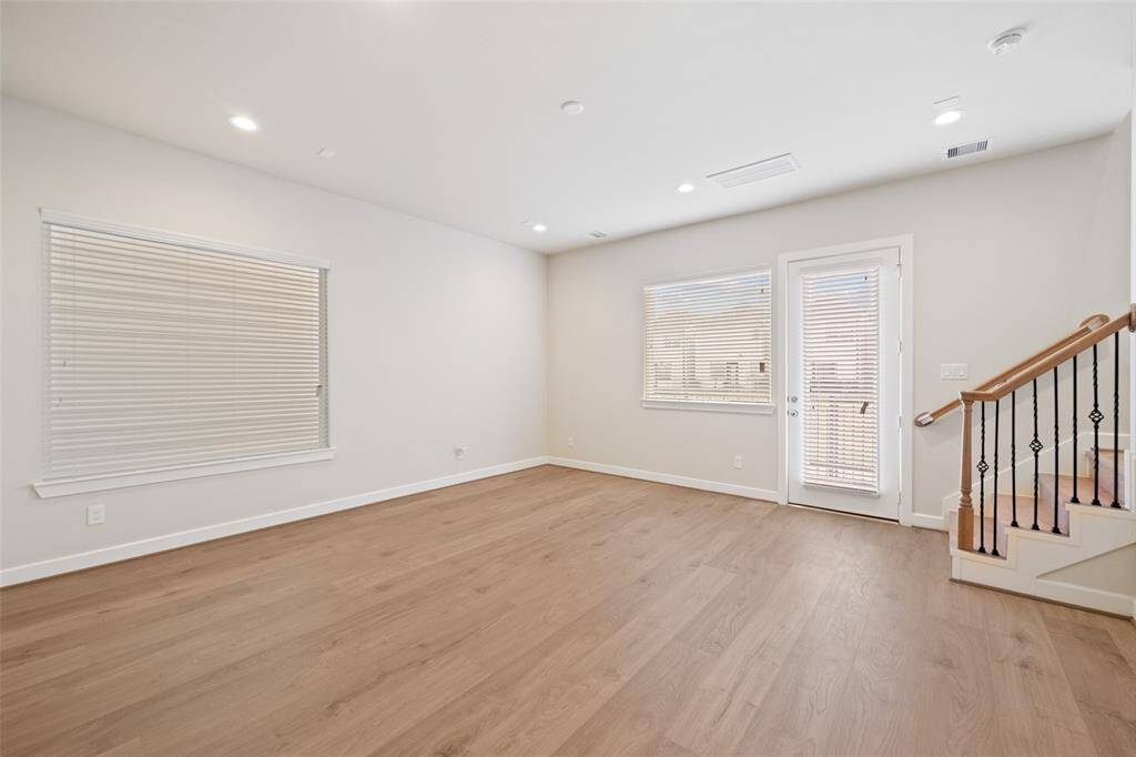 This spacious living area features light wood flooring, large windows with blinds for natural light, and neutral walls. It includes recessed lighting and access to a staircase with elegant railing details.
